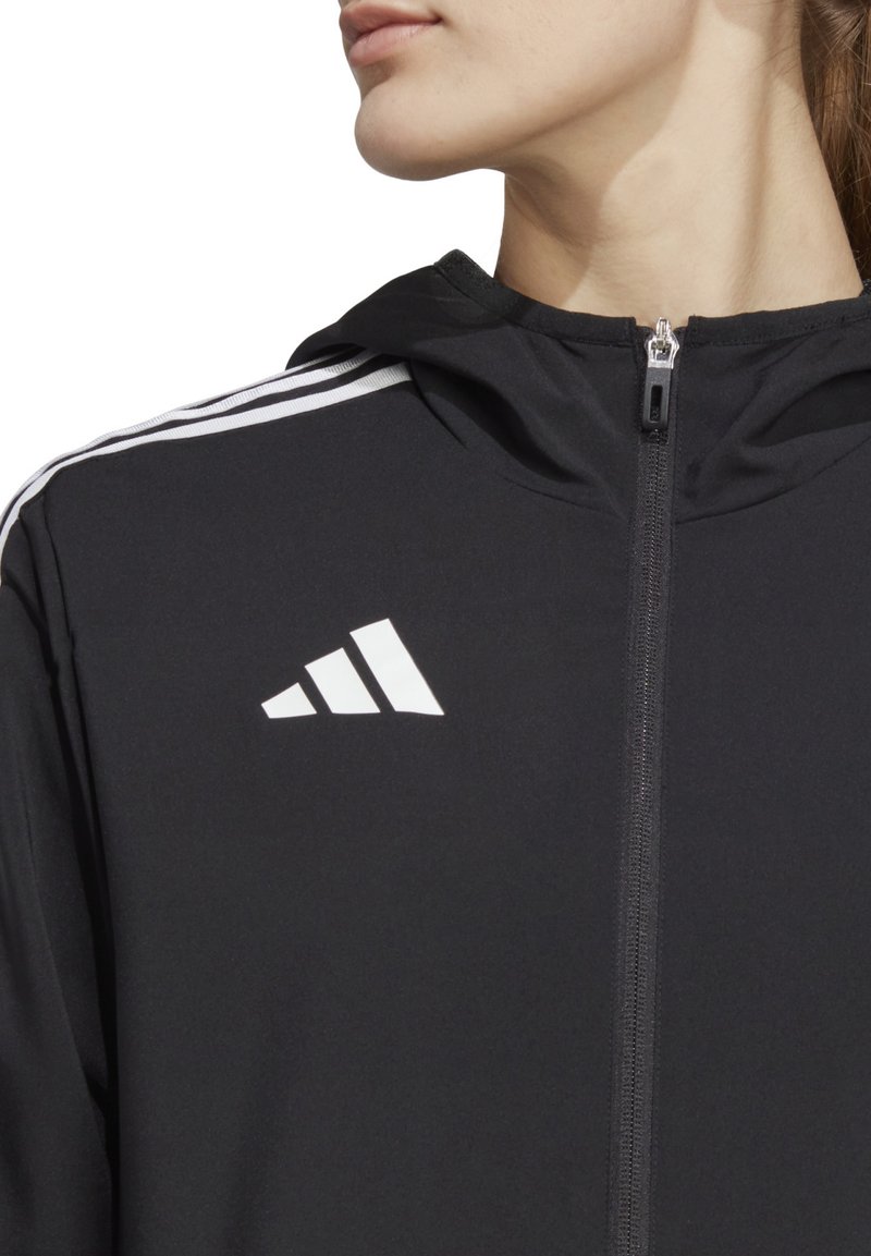 Giacca nera con zip e cappuccio, con logo Adidas bianco e tre strisce bianche sulle spalle. Tessuto dalla texture liscia.
