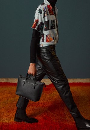 Personne portant un pantalon en cuir noir et une chemise à manches courtes à motifs par-dessus un haut à manches longues noir, tenant un sac à main noir texturé en marchant.