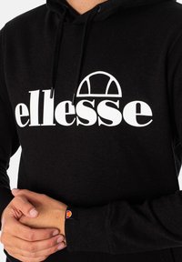Sudadera negra con un gran logo blanco de "ellesse" en la parte frontal. Cuenta con capucha con cordón y puños de canalé. La etiqueta en el puño muestra un logo naranja.