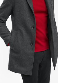 Cappotto di lana grigia misto con design a un petto, due tasche frontali e bottoni neri, indossato su un maglione di lana rossa.