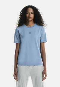 Ljust blå t-shirt med kort ärm, rundad halsringning och en liten logotyp på bröstet. Mjuk tyg med en slät textur.