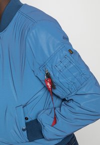 Ljusblå bomberjacka med ribbade mudd och krage. Har en dragkedja på ärmen och diskreta logotypsdetaljer. Slät, nylonmaterial.