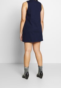 Nainen, jolla on ykköspiirretetty sininen ilmanhiha mekko ja nilkkakorkuiset musta-harmaat käärmeennahkasaappaat, seisoo vaaleanvalkoista taustaa vasten.