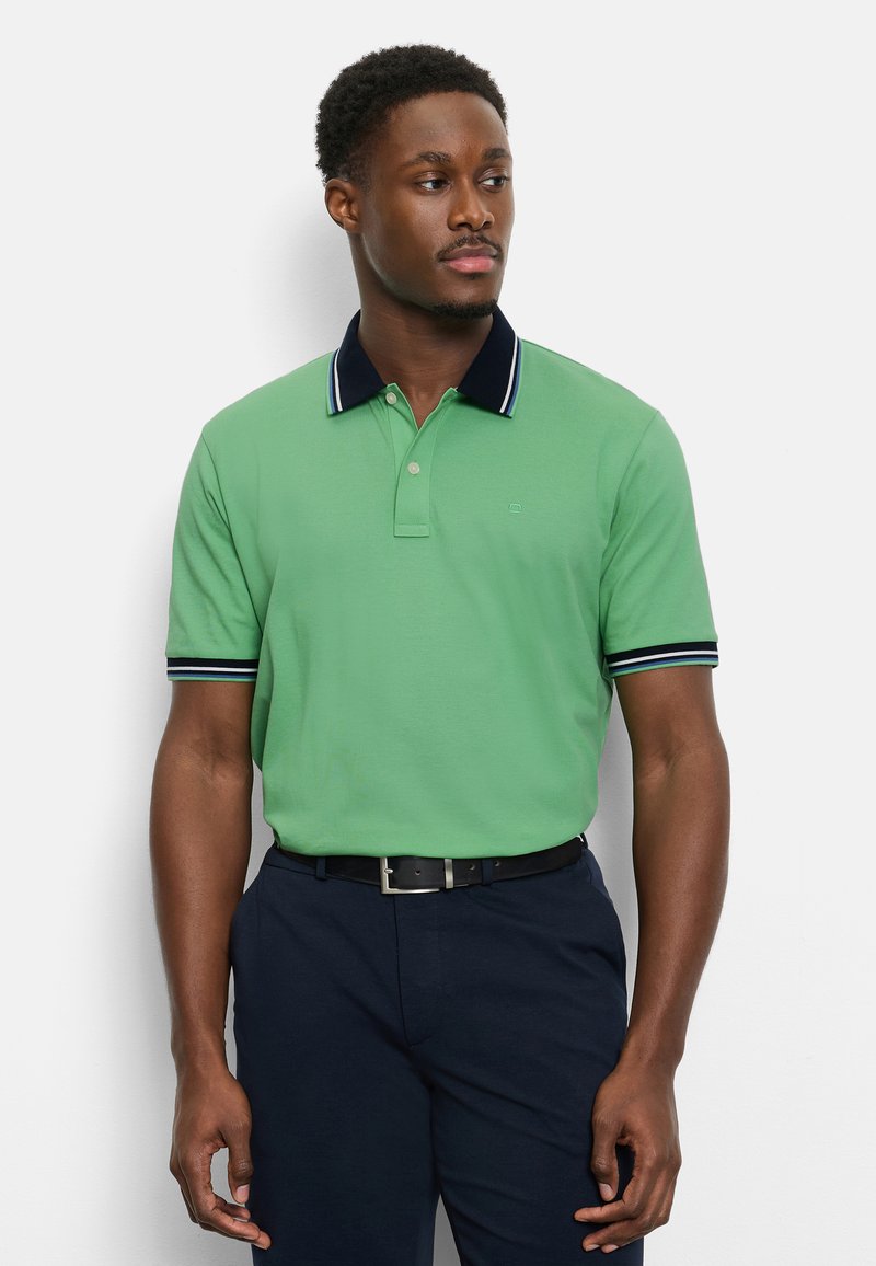 Mann trägt grünes Polo mit kurzen Ärmeln, marineblauem Kragen und Bündchen, marineblaue Hose und schwarzen Gürtel, steht vor weißem Hintergrund.