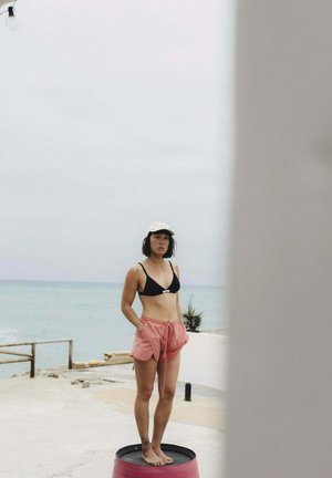 Frau in schwarzem Bikinioberteil und pinken Shorts steht barfuß auf einem runden schwarzen Fass am Meer unter bedecktem Himmel.