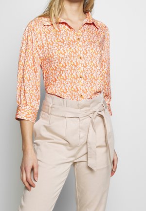 Frau trägt eine orange-weiße, geblümte Bluse mit Knöpfen und hoch taillierte beige Hose mit einem an der Taille gebundenen Gürtel.