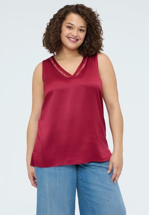 Femme souriante aux cheveux bouclés portant des boucles d'oreilles créoles, un haut sans manches en satin rouge avec des bordures en dentelle, et un jean bleu clair sur un fond uni.