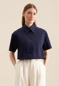 Granatowa koszula z krótkim rękawem i spiczastym kołnierzykiem, zapinana na guziki, uszyta z gładkiego materiału. Białe spodnie z kieszeniami dopełniają stylizację.