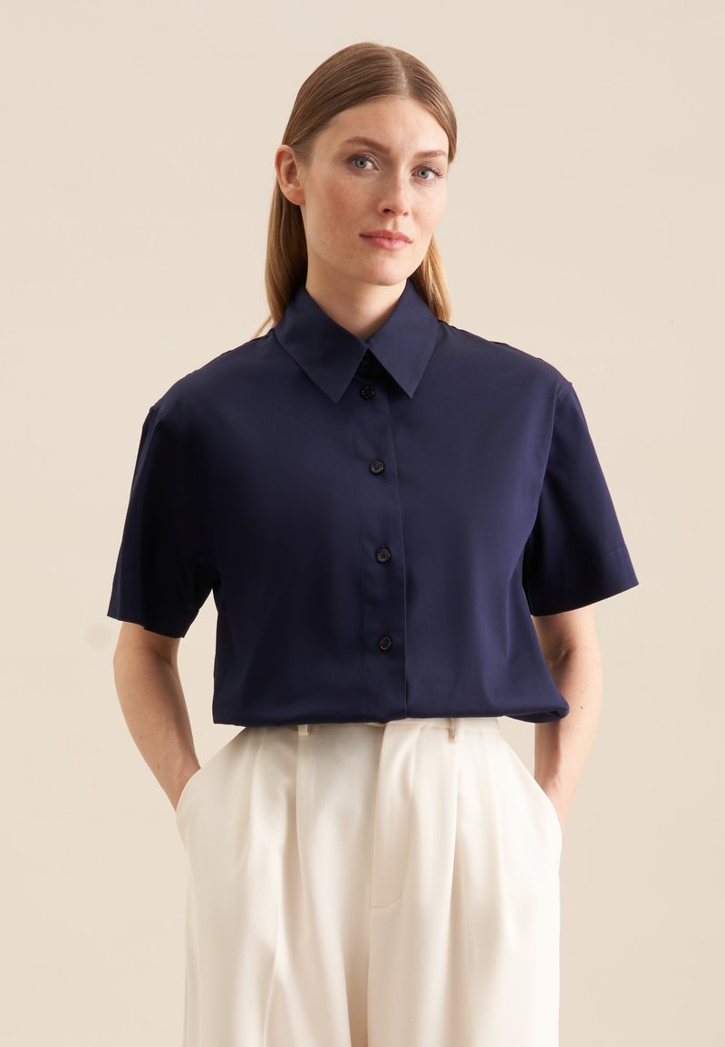 Granatowa koszula z krótkim rękawem i spiczastym kołnierzykiem, zapinana na guziki, uszyta z gładkiego materiału. Białe spodnie z kieszeniami dopełniają stylizację.