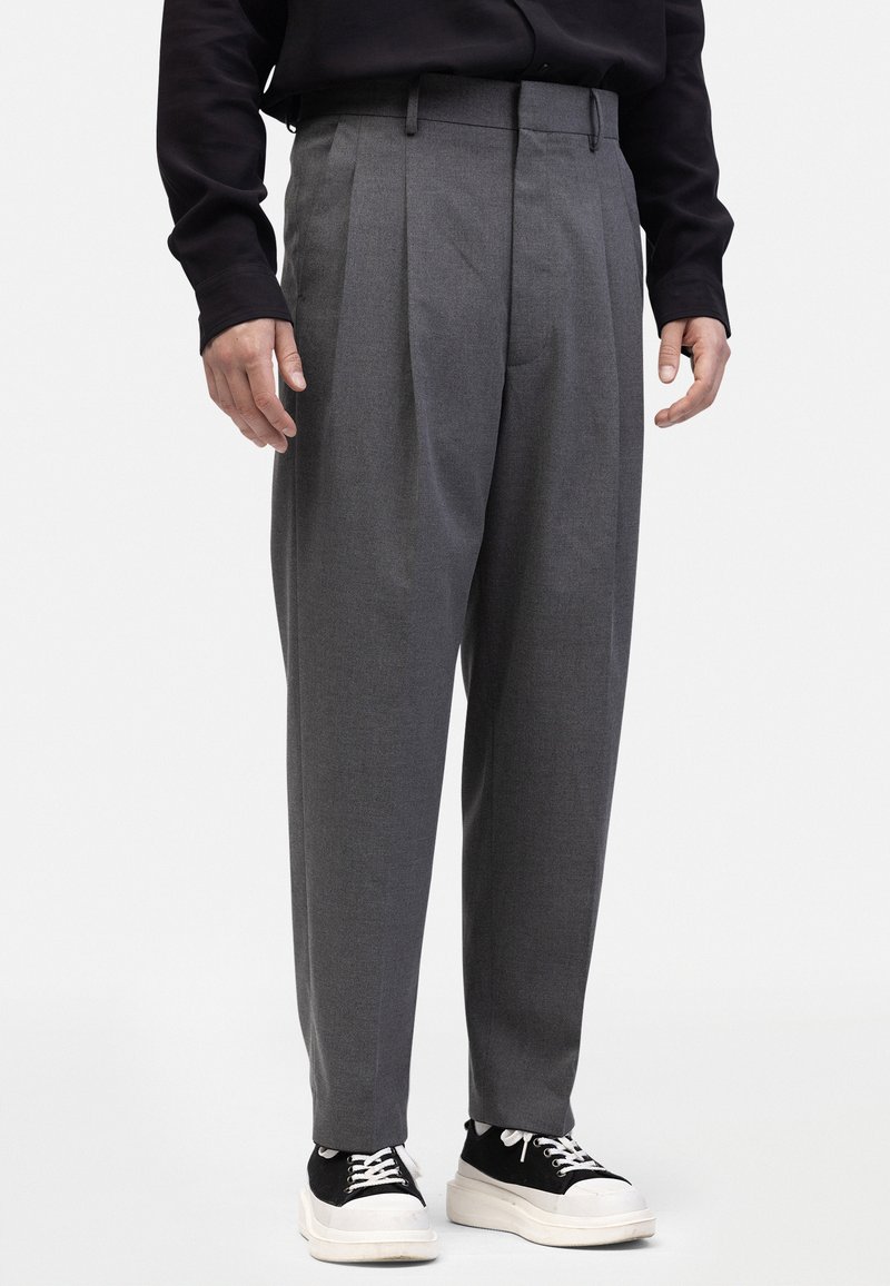 Pantaloni gri plisati, dintr-un material țesut, cu o croială relaxată, cu buzunare laterale și asortați cu adidași negri și albi.