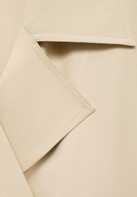 Beige tyg med en slät yta, som har vinklade överlappande flikar och noggrann sömnad. Designen betonar skräddarsydda linjer och geometriska former.