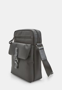 Borsa a tracolla nera con tasca frontale a pattina bloccata da una fibbia, scomparto superiore con zip e tracolla regolabile su uno sfondo bianco.