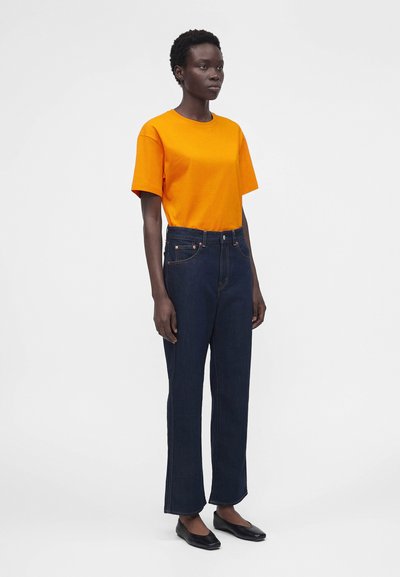 Femme debout portant un t-shirt orange vif à manches courtes, un jean bleu foncé taille haute et des chaussures plates noires sur un fond gris uni.