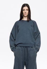 Prešívaná tmavomodrá mikina vo veľkosti oversized, vyrobená z bavlny, s okrúhlym výstrihom, rebrovanými manžetami a vyšitým logom na hrudi.