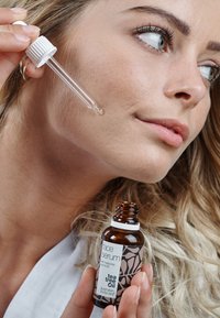 Brun glassflaske med tea tree olje ansiktsserum og hvitt lokk. Klar pipette i hånden, påfører serum på huden. Lysebrunt hår.