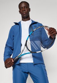 Modrá sportovní bunda se zipem vpředu, bílá košile s límečkem pod tím a modré kalhoty. Drží černou tenisovou raketu s modrými akcenty.