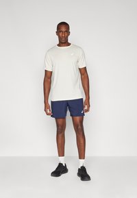 Men's light beige athletic t-shirt with short sleeves, paired with navy shorts. Model wears black sneakers and white socks. Simple logo detail visible.