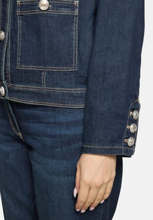 Gros plan sur une veste en jean avec des boutons argentés sur le poignet et la poche, portée avec un jean bleu foncé assorti, main reposant le long du corps.