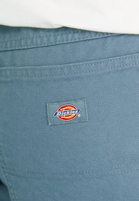 Sininen Dickies-kangas teksturoidulla pinnalla, jossa on suorakulmainen logo-patch punaisella, keltaisella ja sinisellä takana.