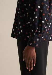 Blusa nera con stampa floreale multicolore, maniche lunghe, polsini con bottone e texture morbida. Abbinata a pantaloni scuri.