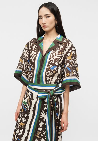 Γυναίκα που φοράει ένα φαρδύ φόρεμα με καφέ, πράσινο, μπλε και λευκό μοτίβο, με ζώνη στη μέση δεμένη και κοντά μανίκια, στέκεται μπροστά σε μια απλή φόντο.