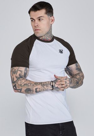 Homme avec des tatouages sur le bras et le cou portant un t-shirt blanc à manches raglan noires, les mains jointes et regardant sur le côté devant un fond uni.