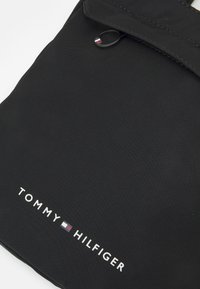 Borsa nera in nylon con chiusura a pattina, anello decorativo e logo "TOMMY HILFIGER" in testo bianco con un emblema colorato.