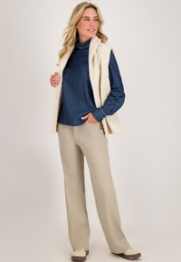 Crème mouwloze vest over een marineblauwe blouse, beige broeken en lichtcrème schoenen. De blouse heeft een hoge hals en knoopaccenten op de mouwen.