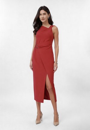 Ženska z dolgimi temnimi lasmi, oblečena v rdečo midi obleko brez rokavov z razporkom na strani in zlatim sponkom, bež salonarji, srebrna zapestnica in uhani.