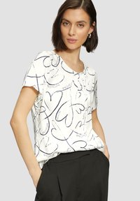 Chemisier blanc avec des motifs de cœurs noirs, à manches courtes, un col rond et une coupe loose, associé à un pantalon noir.