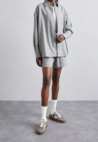 Chemise grise à boutons avec une coupe ample, associée à un short assorti. Le modèle porte des chaussettes blanches et des baskets avec un motif à carreaux et des accents beiges.