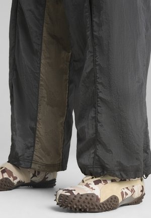 Black and olive patterned pants with a shiny texture; paired with beige camouflaged shoes featuring a spiked sole and secure fit.