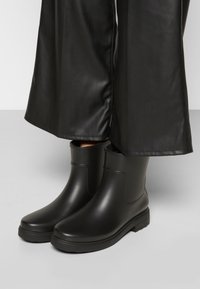 Botas negras hasta el tobillo hechas de goma suave con punta redondeada, tacón bajo y diseño minimalista. Combinadas con pantalones anchos negros.
