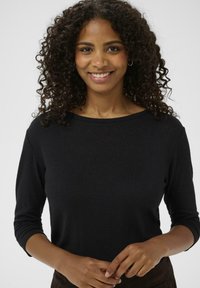 Haut noir à col rond et manches longues en tissu doux, avec un design ajusté et des poignets côtelés, sur un fond neutre.