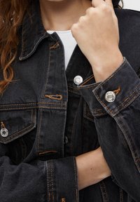 Giacca di jeans nera con taglio classico, dotata di bottoni metallici, cuciture visibili e particolari scoloriti sul tessuto.