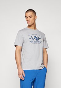Camiseta gris de manga corta con un gráfico de montaña y el texto "Columbia" en azul marino. Corte casual, textura suave, cuello redondo.