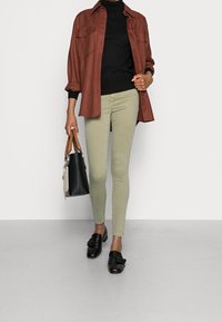 Camisa de gran tamaño en color óxido, suéter de cuello alto negro, jeans ajustados en verde claro, mocasines negros con hebilla y un bolso negro con asas marrones.