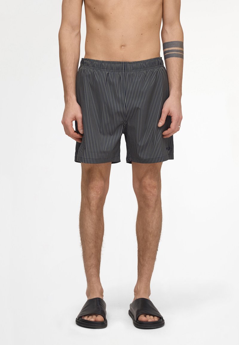 Personne torse nu portant des sandales slide noires et un short noir avec de fines rayures verticales grises, debout sur un fond blanc.