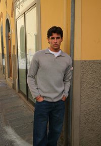 Grå sweater med krave, båret over en hvid skjorte, parret med blå jeans. De står foran en tekstureret væg i et bymiljø.