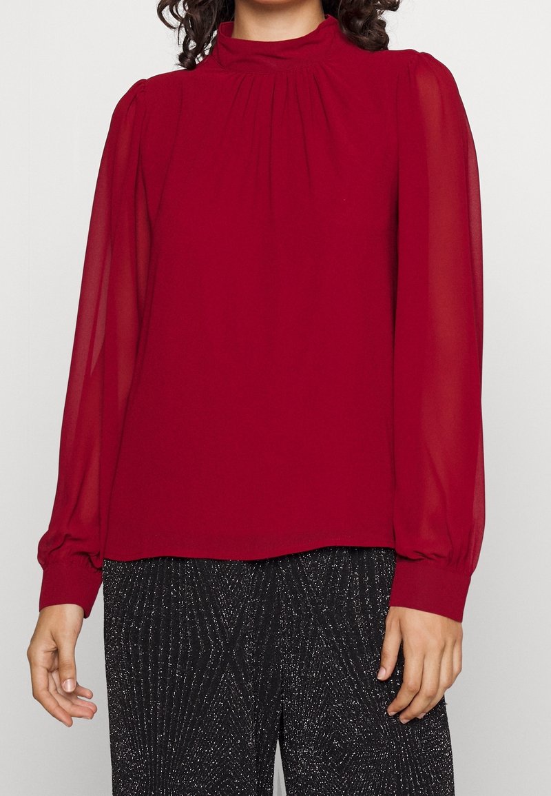 Blouse rouge à col montant avec des manches longues douces et semi-transparentes ; présente des détails froncés au niveau du cou et des poignets. Assortie à un pantalon noir pailleté.
