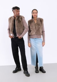 Două modele stau una lângă alta. Ambele poartă veste din blană sintetică de culoare maronie; una are un pulover bej cu dungi dedesubt, iar cealaltă poartă un top cu model. Modelul masculin are pantaloni negri și pantofi negri, iar modelul feminin poartă o fustă din denim albastru deschis, cu o deschidere frontală, și cizme negre până la gleznă cu accente argintii.