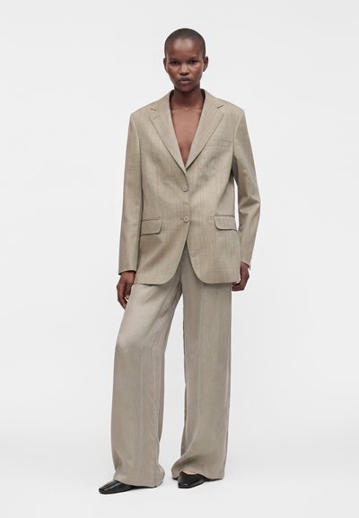 Personne portant un costume beige à fines rayures oversize avec un pantalon à jambes larges et des chaussures noires, debout devant un fond blanc uni.