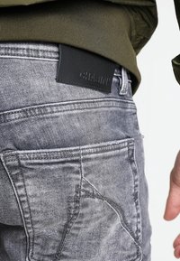 Jeans de mezclilla gris con un acabado texturizado, que cuentan con una etiqueta de marca de cuero negro en la cintura y un gran detalle de bolsillo lateral.