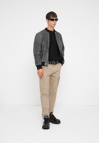 Chaqueta bomber gris con acentos negros, llevada sobre un suéter negro y pantalones cargo beige. Botas negras de suela gruesa, gafas de sol, fondo neutro.