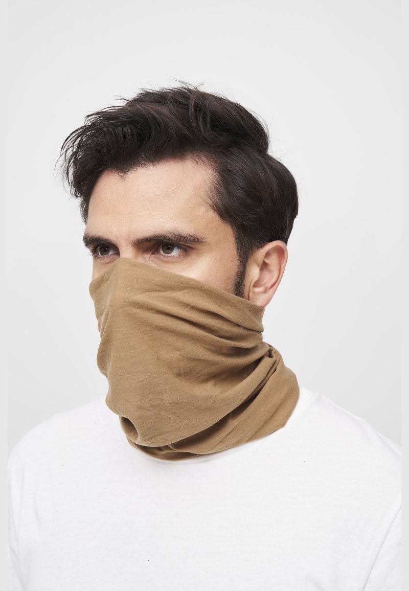 Hombre de cabello oscuro que lleva un braga para el cuello marrón cubriendo la nariz y la boca, vestido con camisa blanca, sobre un fondo liso.