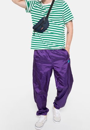 Person trägt ein grün-weiß gestreiftes T-Shirt, glänzende lila Hose, weiße Turnschuhe und eine schwarze Umhängetasche.