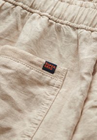 Tessuto beige chiaro con una finitura testurizzata, dotato di una vita elasticizzata e una tasca a patta. È visibile un'etichetta "Superdry" blu e arancione.