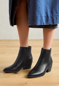 Bottines en cuir noir avec un bout pointu, un petit talon carré et une fermeture éclair sur le côté, portées avec une jupe en denim, sur un sol en bois.