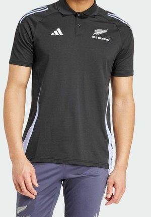 Homme portant un polo noir Adidas All Blacks avec des bandes blanches et un pantalon de sport gris avec logo sur la cuisse, sur fond uni.