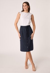 Top bianco senza maniche con spalle arricciate, abbinato a una gonna blu navy fino al ginocchio con vita regolabile e chiusura a bottoni.